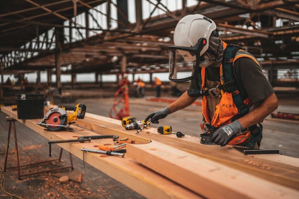 A builder in a hard hat, visor and orange vest with a power tool in hand, working on timber beams on a building site