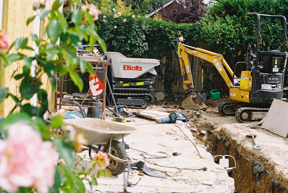 A yellow mini digger and a tracked dumper working in a garden trench, with a wheelbarrow and tools in the foreground