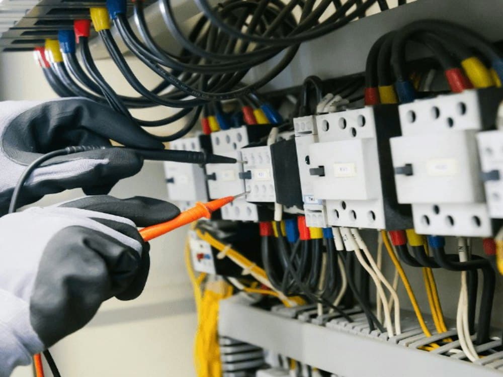 An electrician in gloves testing a panel of contactors and wiring with a multimeter probe