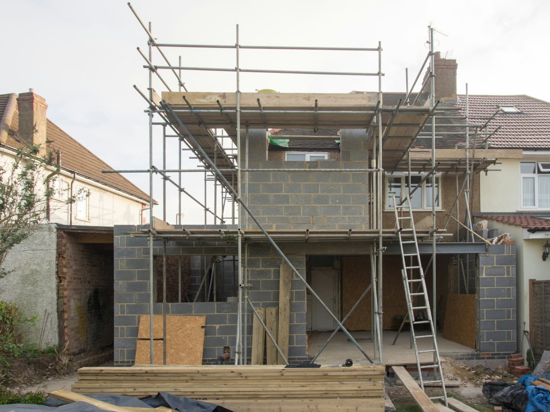 A semi-detached house with a brick extension going up inside scaffolding, a ladder against it and timber stacked in the garden