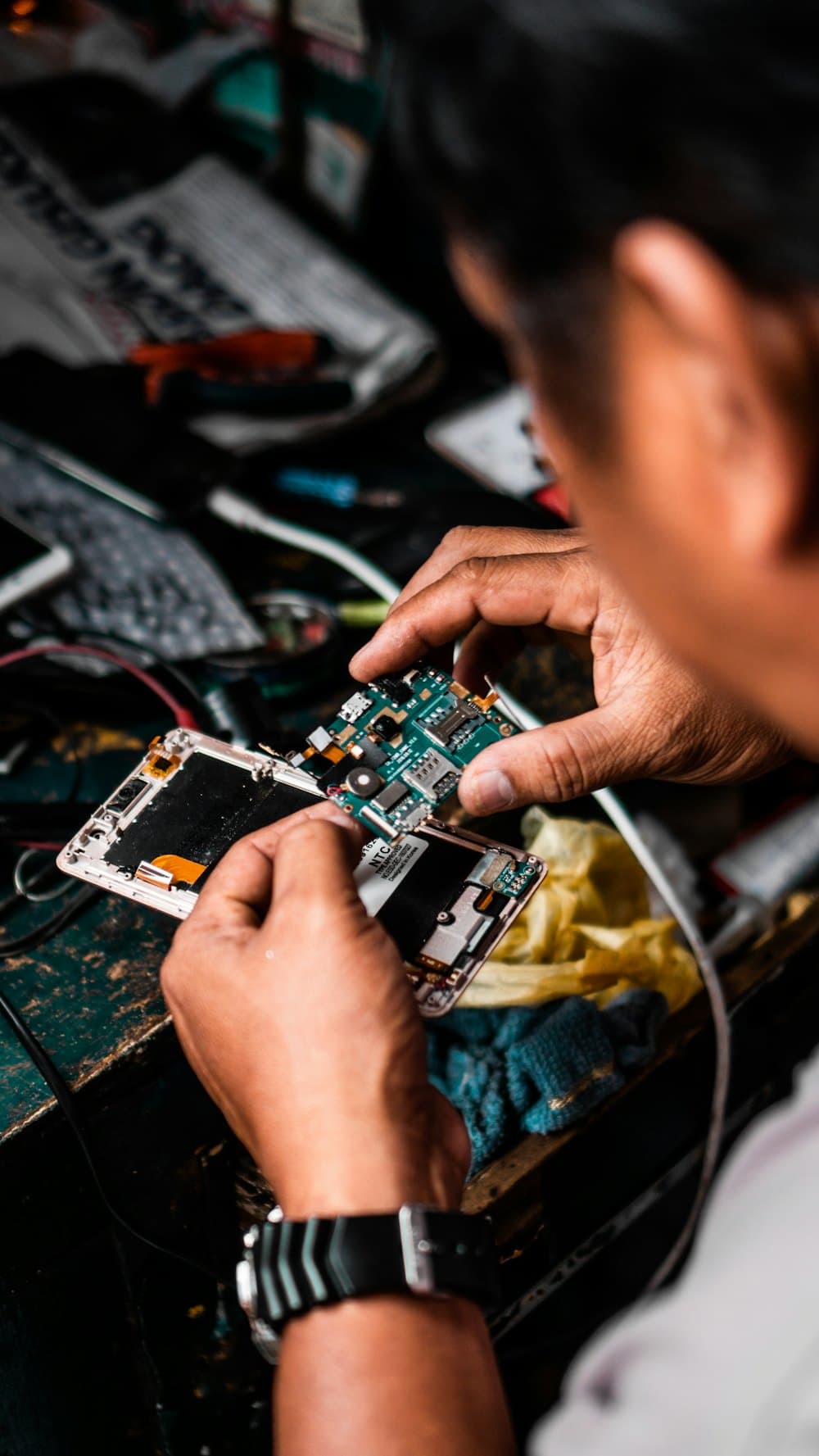 A technician repairing a smartphone at a cluttered bench, holding the open handset and its circuit board